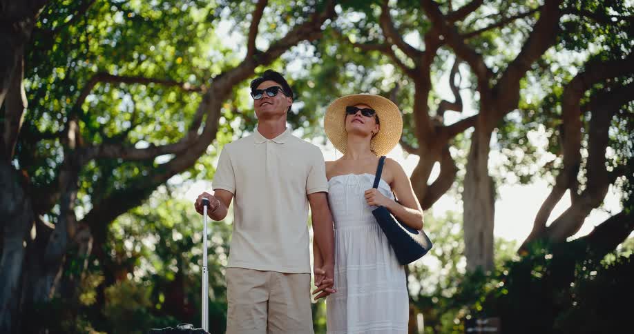 Happy attractive couple holding hands with their roller bag arrives at a tropical resort for their honeymoon vacation