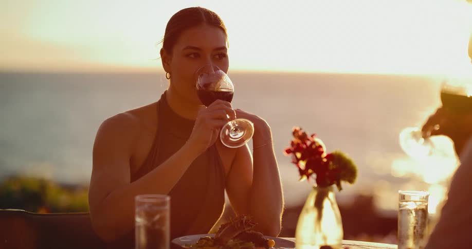 A smiling couple toasts with red wine during a romantic dinner as the sun sets over the ocean in slow motion