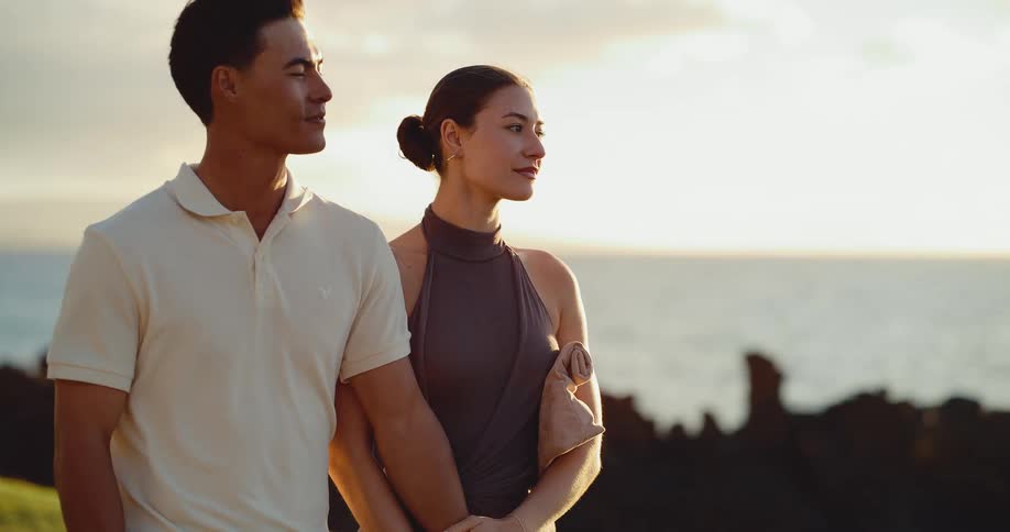 A well-dressed couple walks hand in hand along a coastal path at sunset, enjoying the romantic ocean view