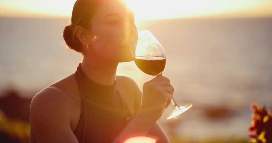 An attractive woman sips a glass of red wine and smiles during a beautiful sunset dinner by the ocean