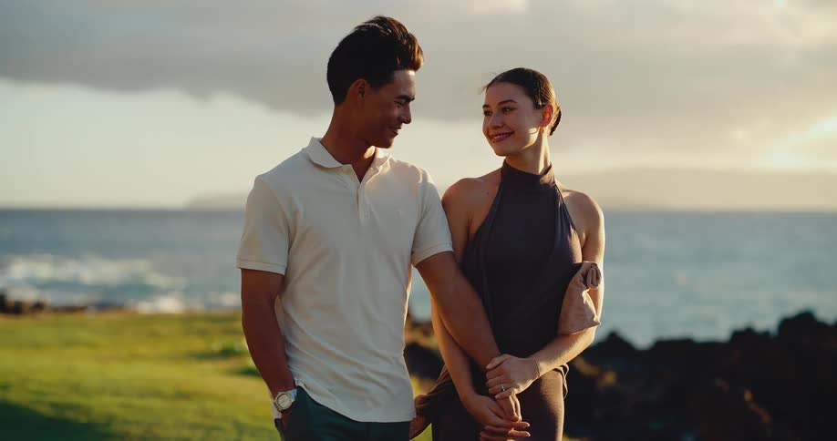 A romantic couple holds hands while walking along a grassy coast and enjoying the beautiful sunset over the ocean