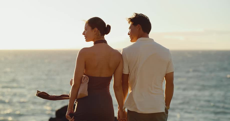 A romantic couple holds hands while standing on a rocky coastline, gazing at the beautiful ocean sunset