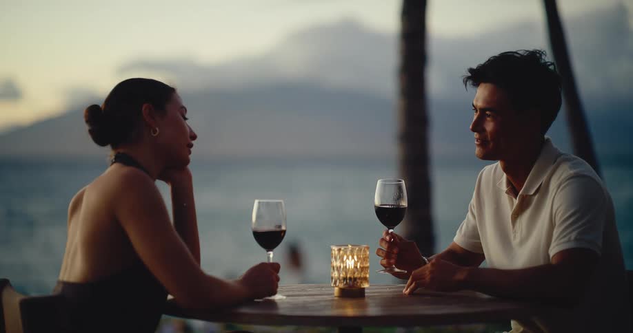A romantic couple enjoys an intimate conversation while drinking red wine at a beautiful tropical resort with an ocean view at sunset