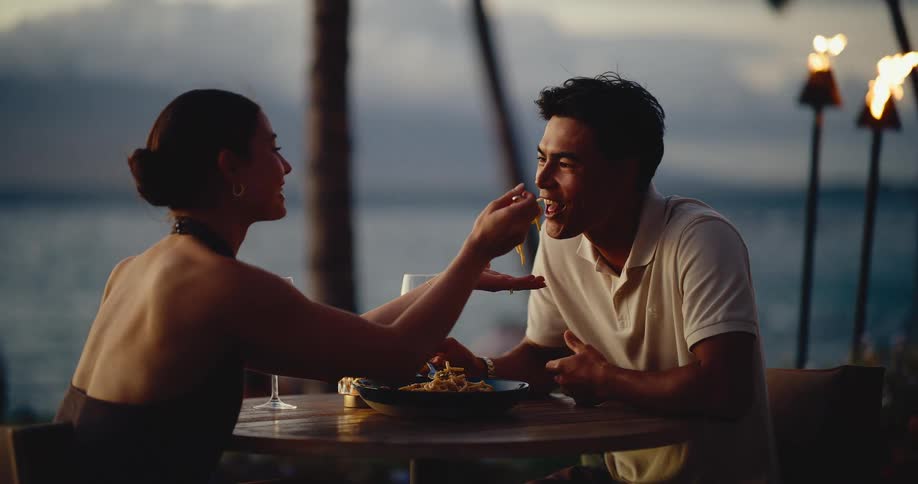 A woman playfully feeds her partner pasta during a romantic beachside dinner at dusk in slow motion