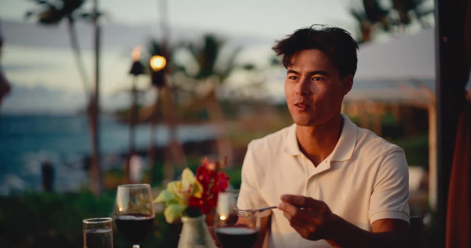 A happy man enjoys a delicious meal during a romantic dinner date with his partner at a beautiful tropical resort at sunset