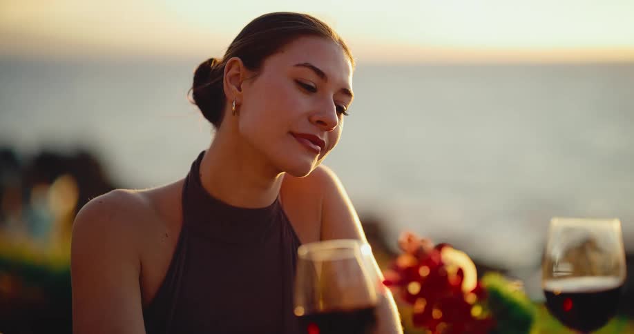 An over-the-shoulder view shows an attractive young woman smiling lovingly at her date before they toast with glasses of red wine during a romantic seaside dinner at sunset