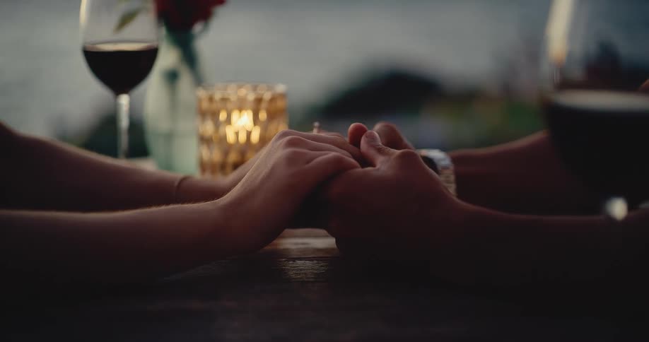 A close-up of a couple holding hands across a romantic, candlelit dinner table at dusk