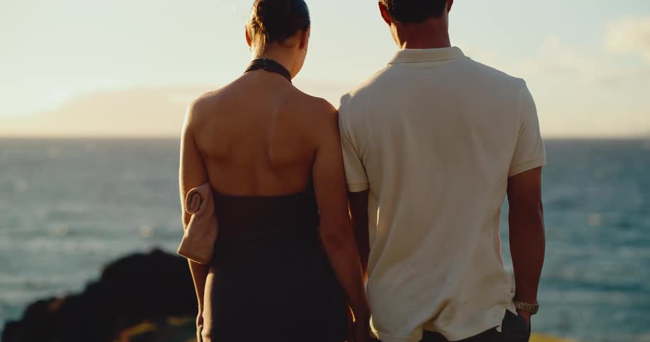 A loving couple holds hands from behind while looking out at the ocean during a beautiful golden sunset