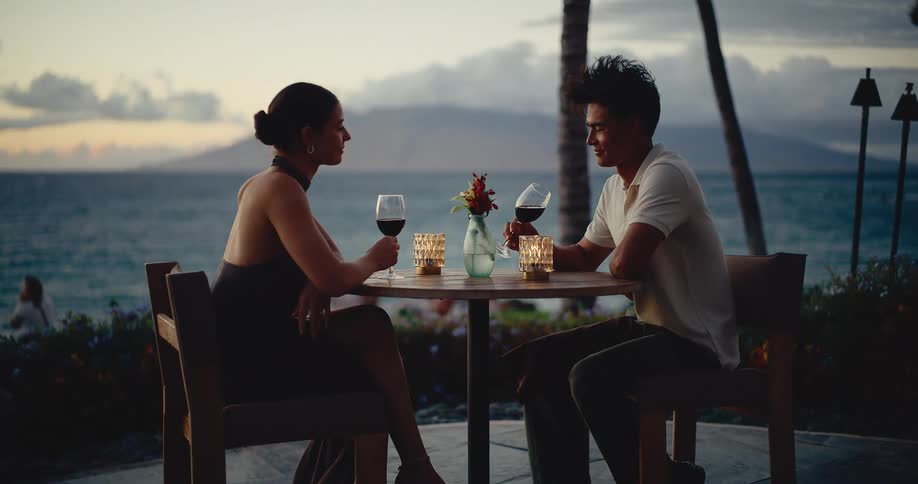 A stylish young couple enjoys a romantic candlelit dinner with red wine at an oceanfront table at dusk