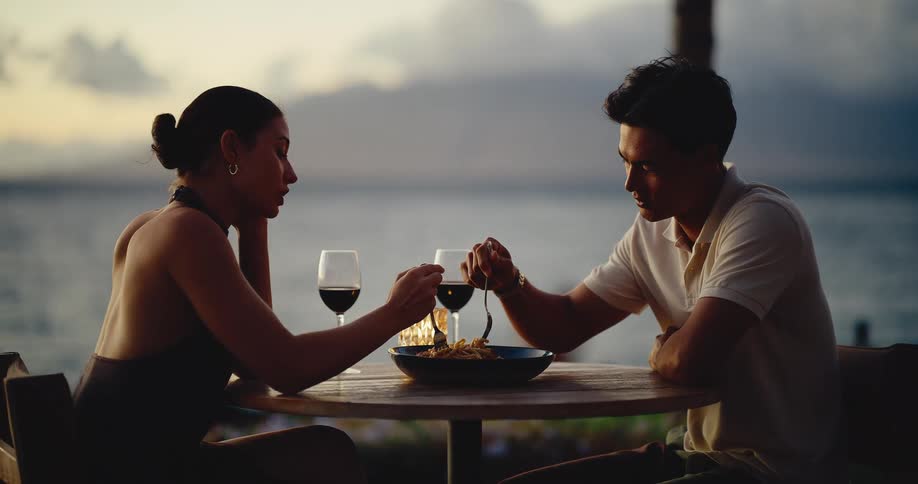 A young couple shares a romantic pasta dinner and red wine at an outdoor table overlooking the ocean at sunset
