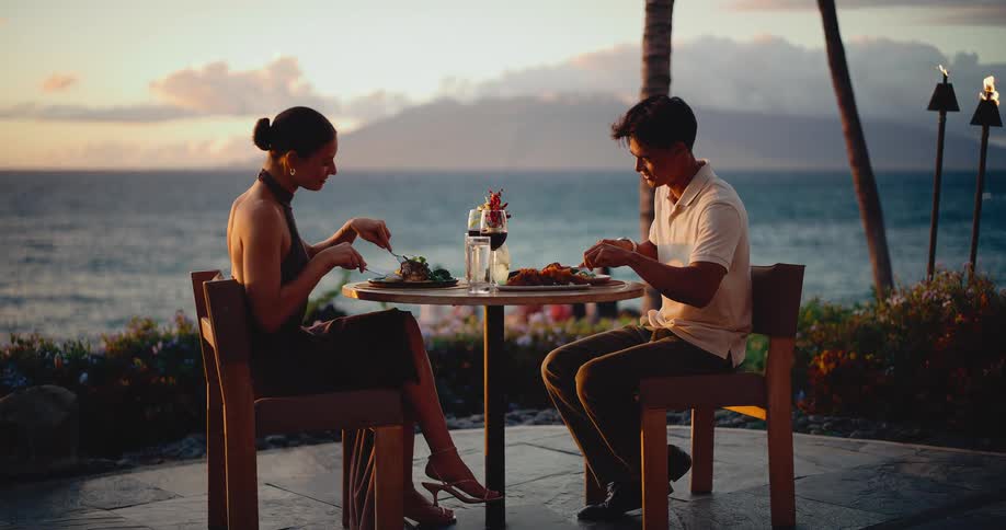 A slow zoom reveals an elegant couple enjoying a romantic dinner for two with a stunning ocean view at sunset