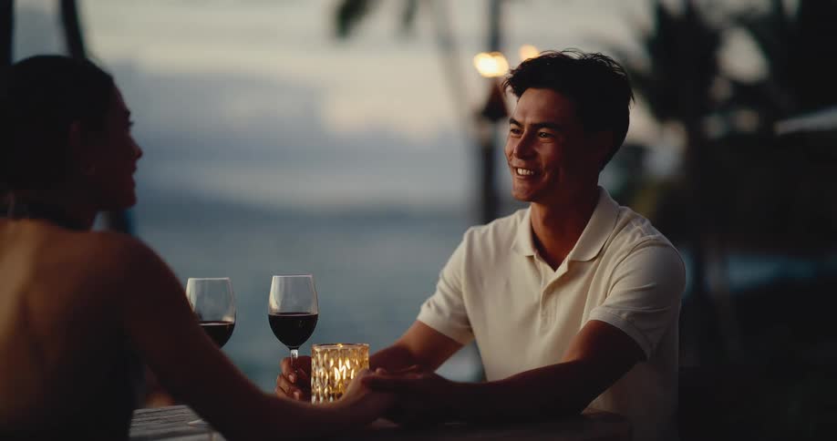 An attractive couple holds hands and smiles lovingly during a romantic candlelit dinner with red wine at a beautiful tropical resort