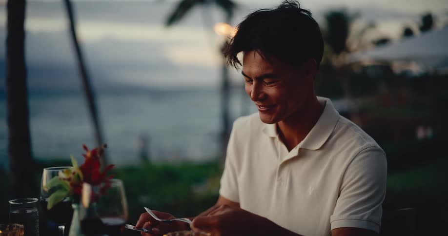 A handsome young man smiles and enjoys a romantic candlelit dinner at a beautiful beachfront restaurant at sunset
