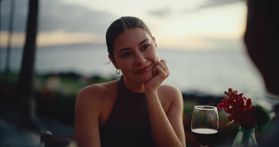 A beautiful young woman smiles at her partner during a romantic sunset dinner with an ocean view