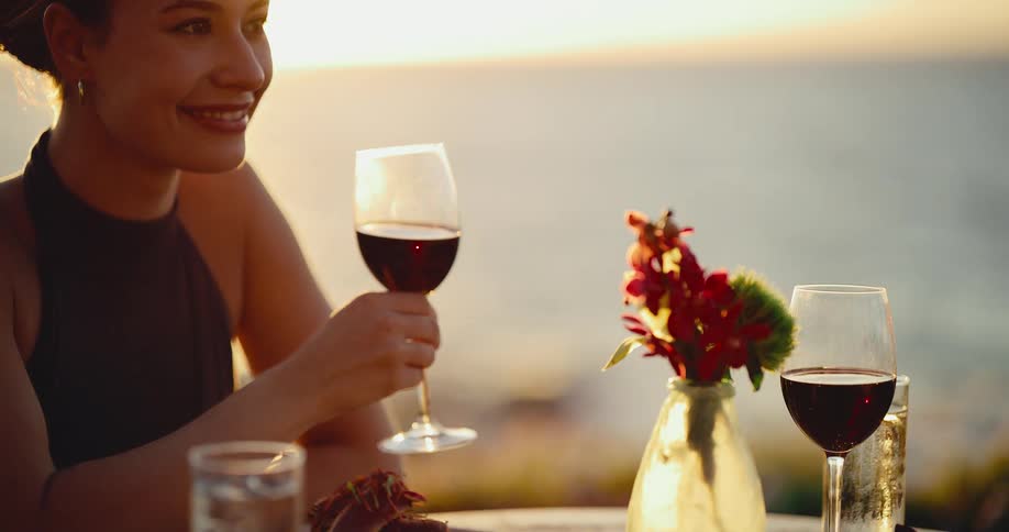 Beautiful young woman sips red wine at a romantic dinner by the ocean as the sun sets in slow motion