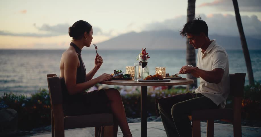 Slow zoom-in on a romantic couple enjoying an elegant dinner with wine at a table overlooking the ocean at sunset