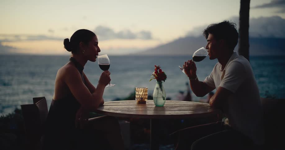A young couple toasts and sips red wine during a romantic dinner date overlooking the ocean at sunset