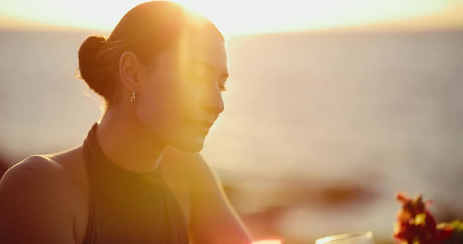 A beautiful woman drinks a glass of red wine and enjoys a romantic dinner overlooking the ocean at sunset