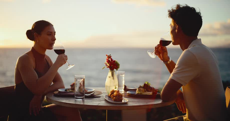 A young, attractive couple toasts with red wine during a romantic sunset dinner at a luxurious oceanside restaurant