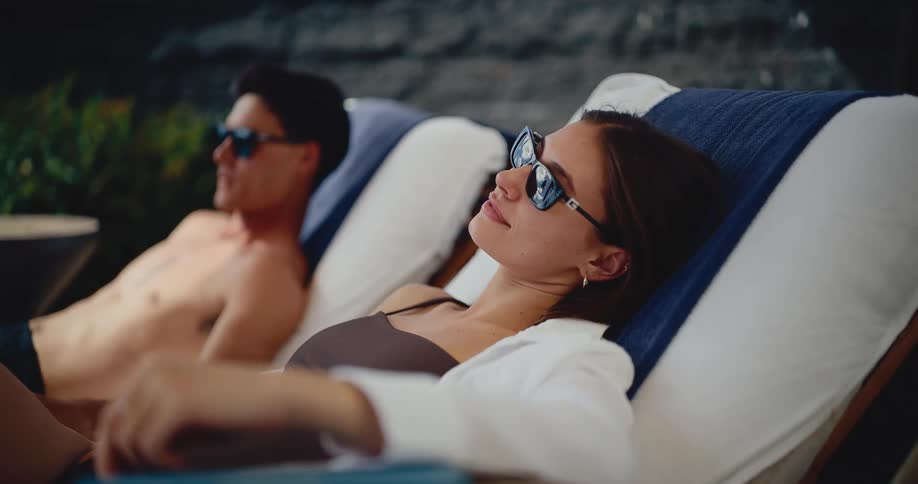 A relaxed young couple in sunglasses sunbathes on lounge chairs at a luxury resort
