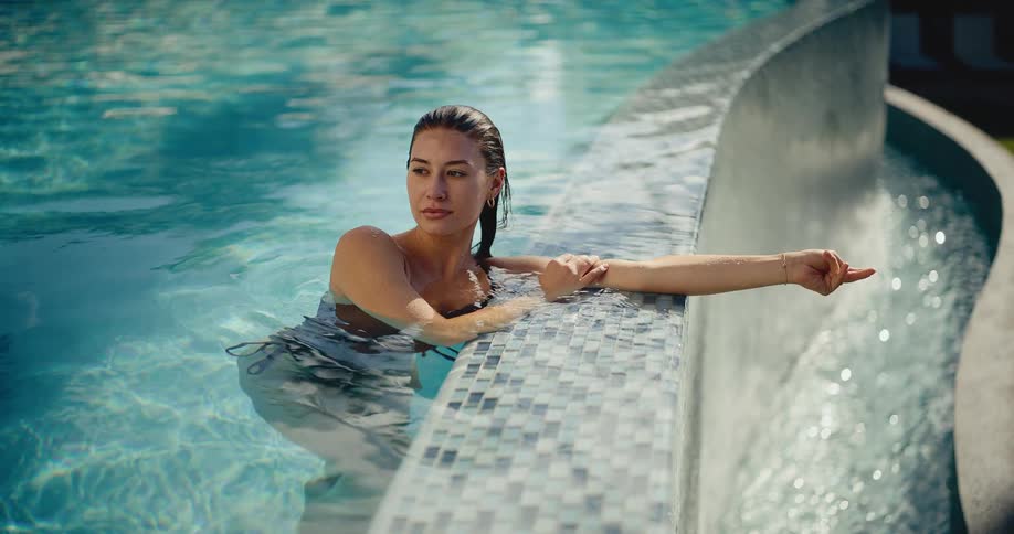 A beautiful young woman with wet hair relaxes by the tiled edge of a sunlit swimming pool