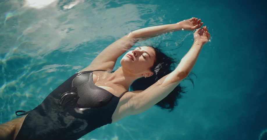 A serene woman in a black swimsuit peacefully floats on her back in the calm, blue water of a swimming pool on a sunny day