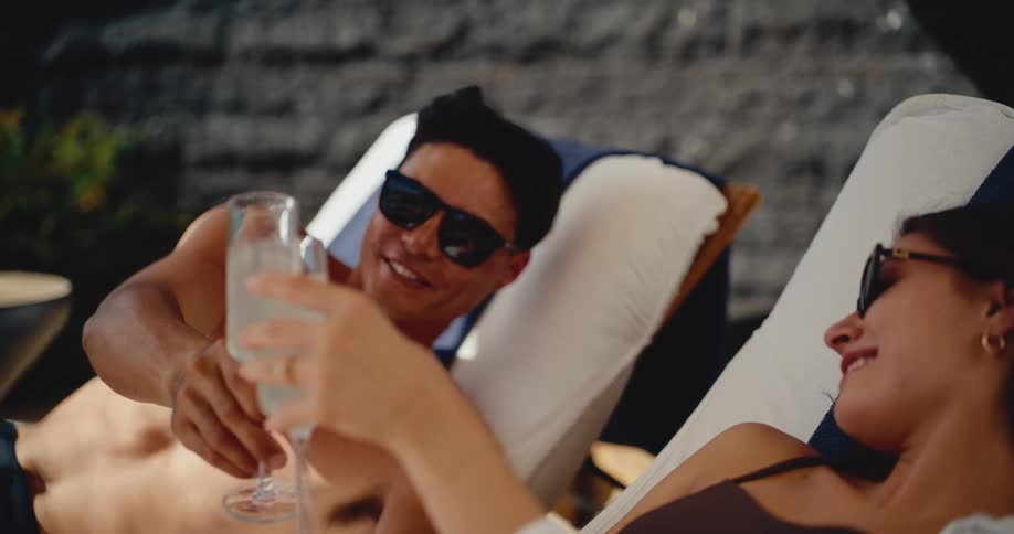 An attractive couple wearing sunglasses toasts with champagne while relaxing on lounge chairs at a luxury resort