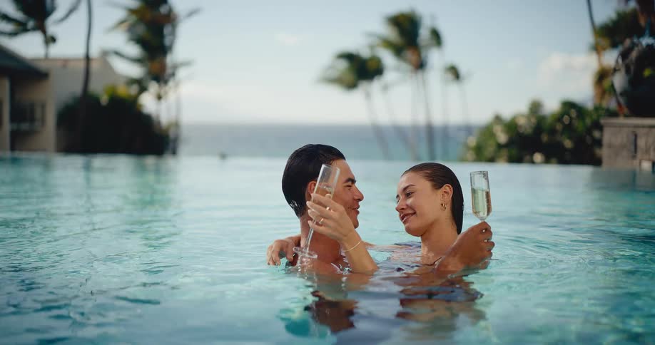 A romantic couple embraces and toasts with champagne glasses while relaxing in a luxury infinity pool at a tropical resort