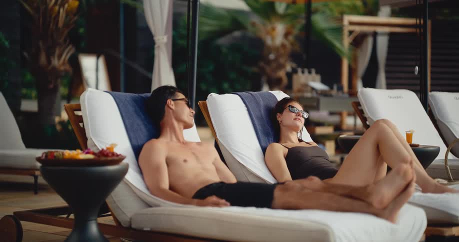 A young, attractive couple relaxes on lounge chairs by a luxurious pool, sunbathing and enjoying their tropical vacation