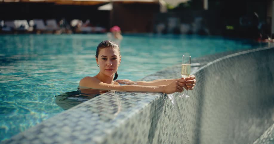 An elegant woman with wet hair relaxes in a luxury resort swimming pool holding a glass of sparkling champagne