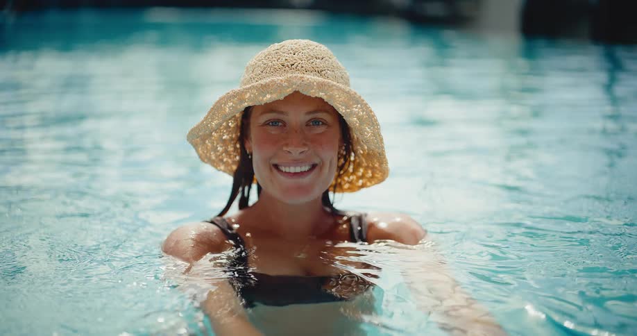 A relaxed woman in a straw hat smiles as she enjoys the calm water of a swimming pool in slow motion