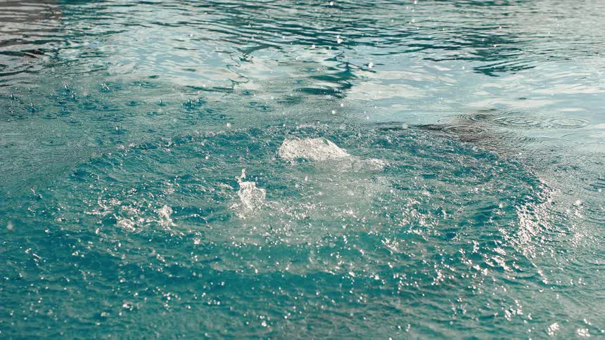 Raindrops and bubbling jets disturb the tranquil surface of a turquoise pool in slow motion