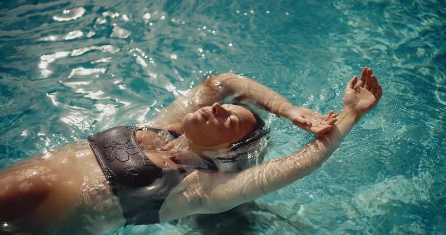 A serene woman in a bikini floats weightlessly in a sun-dappled swimming pool in slow motion