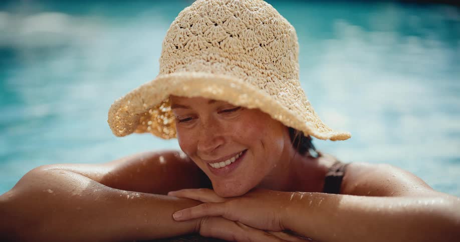 A beautiful woman with freckles and a sun hat smiles while resting on the edge of a sun-dappled swimming pool