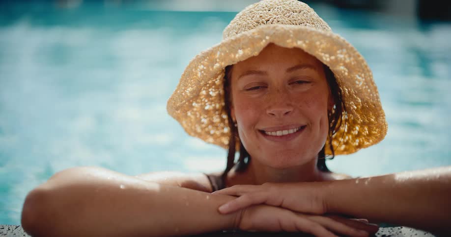 A radiant woman in a straw hat smiles contentedly while relaxing at the edge of a sunlit swimming pool