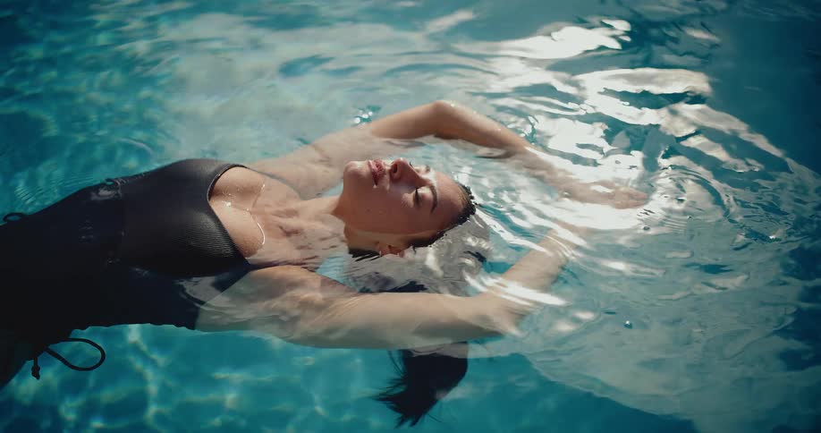A serene woman in a black swimsuit floats peacefully on her back in a sunlit swimming pool