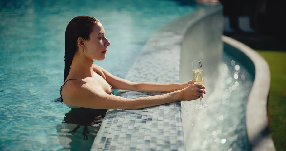 A beautiful woman in a black bikini relaxes in a luxurious swimming pool, holding a glass of champagne and enjoying the sun