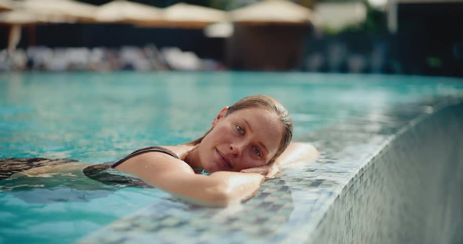 A serene woman with wet hair leans on the tiled edge of a resort swimming pool, closing her eyes in peaceful relaxation