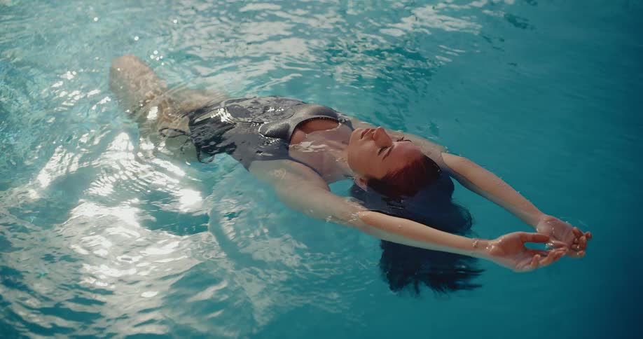 A serene woman in a swimsuit peacefully floats on her back in the clear blue water of a sunlit swimming pool