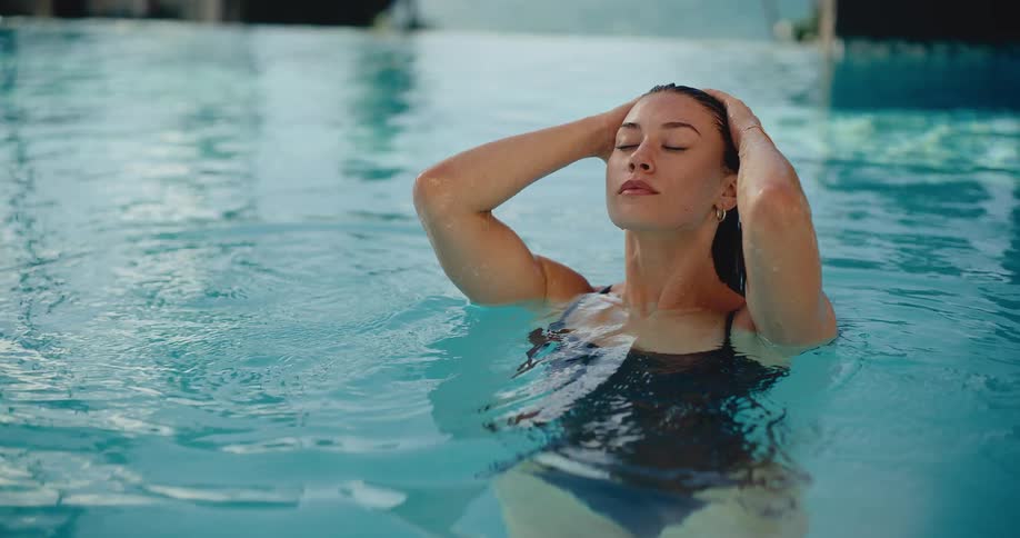 A beautiful young woman in a swimsuit relaxes peacefully in a swimming pool, running her hands through her wet hair in slow motion