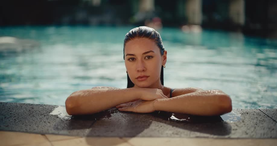 A serene woman with wet hair emerges from the tranquil waters of a swimming pool, resting on the edge before fixing a direct gaze at the camera