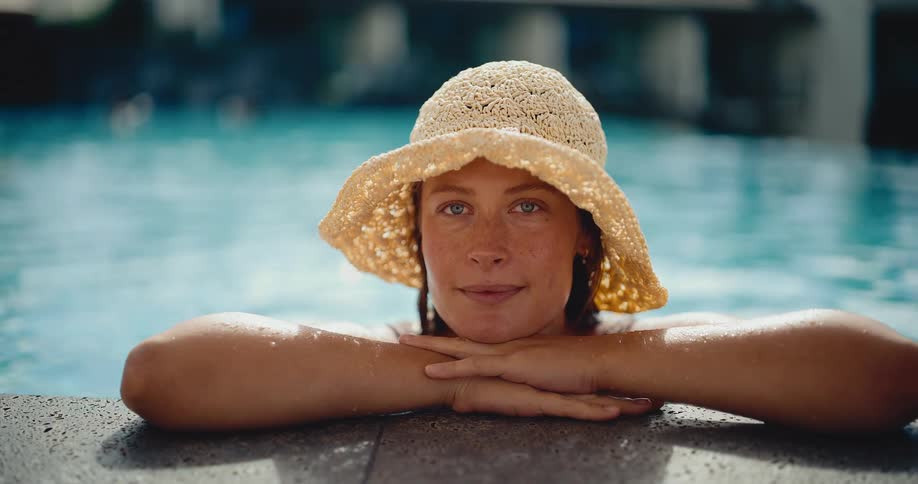A serene woman in a straw hat relaxes at the edge of a sunlit swimming pool, looking peacefully at the camera