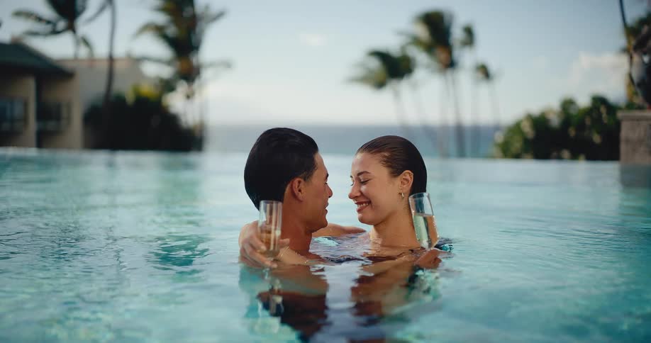 An attractive couple romantically embraces and holds champagne glasses while relaxing in a luxury resort swimming pool