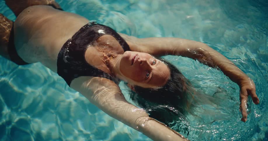 A serene pregnant woman in a bikini floats on her back in a crystal clear swimming pool, looking up at the camera