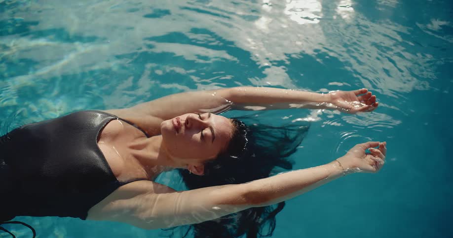 A serene woman in a black swimsuit floats peacefully on her back in a sun-drenched swimming pool