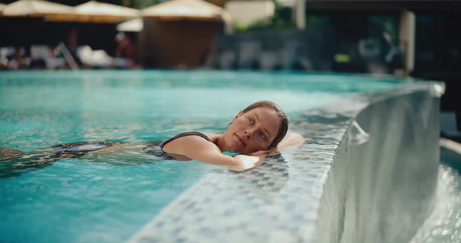 A serene woman in a swimsuit relaxes at the edge of a luxury resort pool in slow motion, opening her eyes to look at the camera