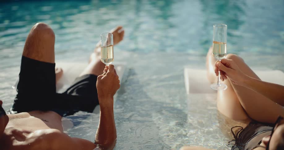 A couple makes a celebratory toast with champagne glasses while relaxing on loungers in a luxurious swimming pool in slow motion