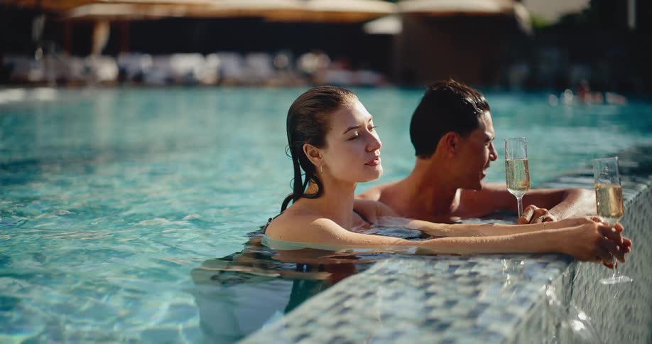An attractive young couple enjoys a romantic and luxurious vacation, relaxing in a resort pool with glasses of champagne