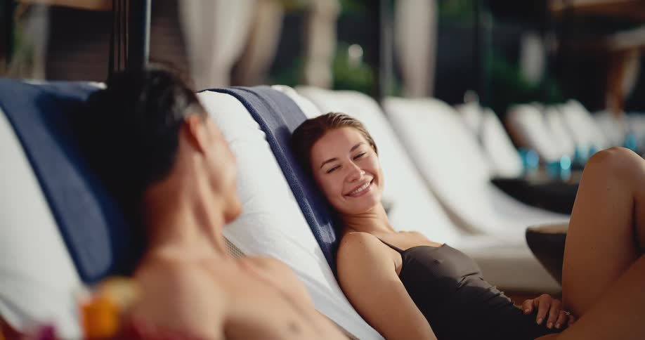A beautiful woman in a black swimsuit smiles at her partner as they relax on poolside lounge chairs