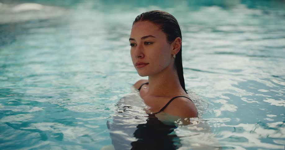 A beautiful young woman with wet hair looks at the camera with a gentle smile while relaxing in the tranquil waters of a swimming pool